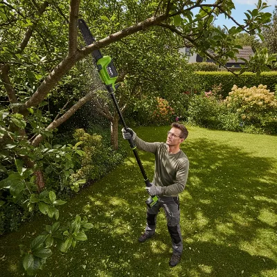 An EGO Power Plus Cordless Pole Saw extending to trim high branches from a tree in a residential garden, with an operator safely on the ground